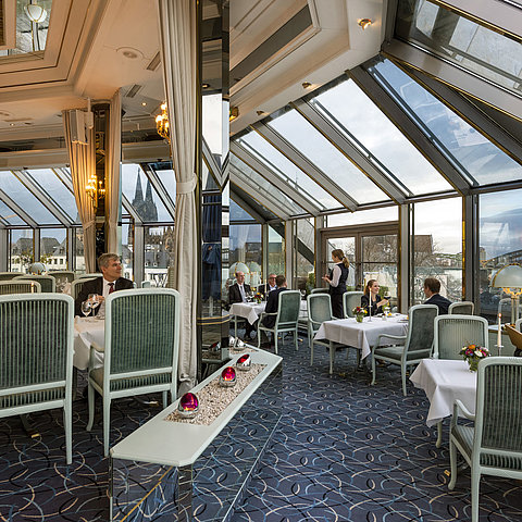 Ristorante elegante con tetto in vetro con vista sul duomo di Colonia e sul ponte Hohenzollern, ospiti che gustano cibo raffinato.