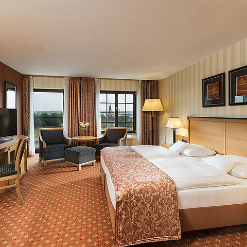 Camera superior al Maritim Hotel Dresden con letto matrimoniale elegante, scrivania e area salotto con vista sulla città.
