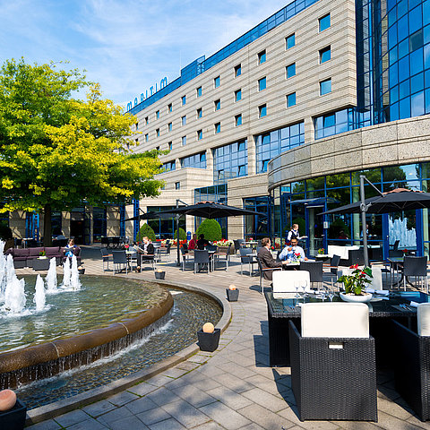 Terrazza dell'hotel Maritim Bonn con aree salotto, fontana e ospiti che cenano e si rilassano all'aperto.