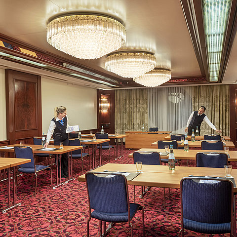 Salone Bonn al Maritim Colonia, il personale prepara la sala con tavoli, bottiglie d'acqua e bicchieri per un evento.