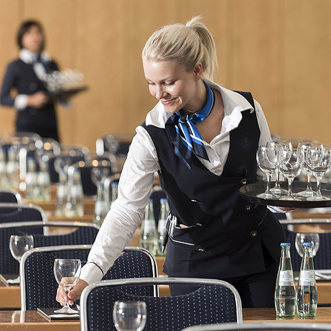 Une employée de serviceprépare une salle de conférence, portant un plateau de verres et disposant des bouteilles d'eau.