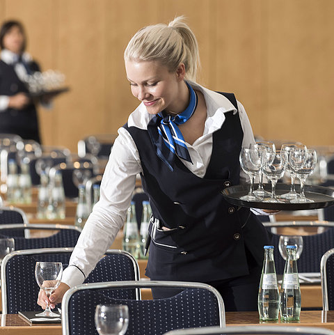 Une employée de serviceprépare une salle de conférence, portant un plateau de verres et disposant des bouteilles d'eau.
