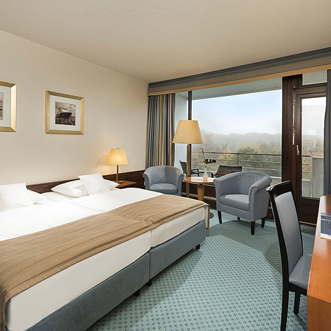 Camera classica del Maritim Hotel Kiel con ampio letto matrimoniale, area salotto, scrivania e balcone con vista sul verde.